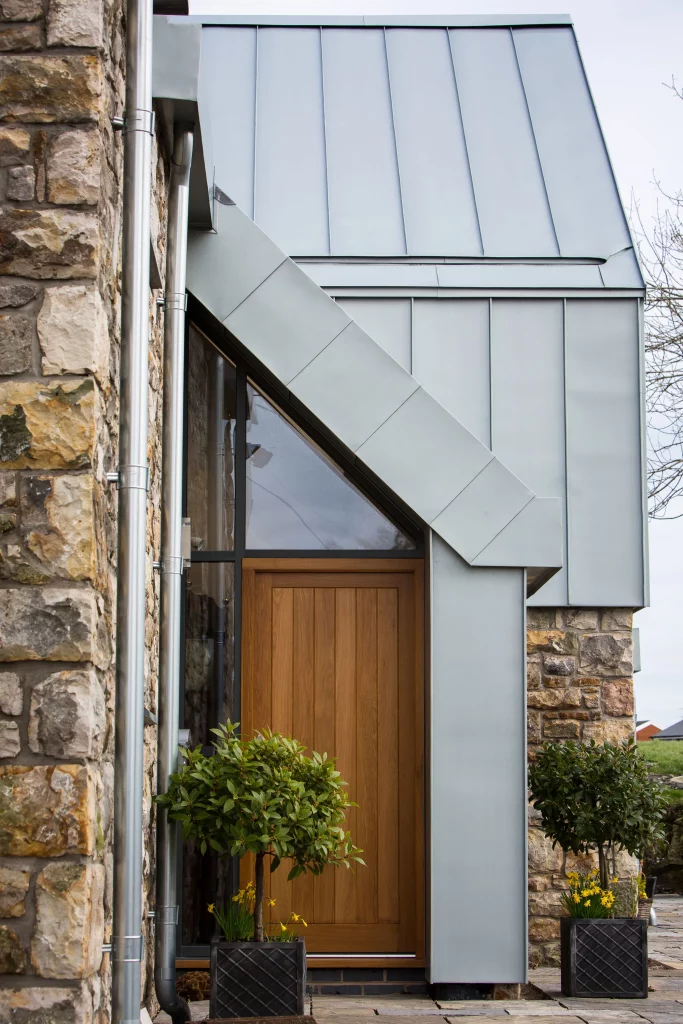 Modern house entrance with wooden door, zinc-clad angled canopy, stone walls and potted topiary with yellow flowers