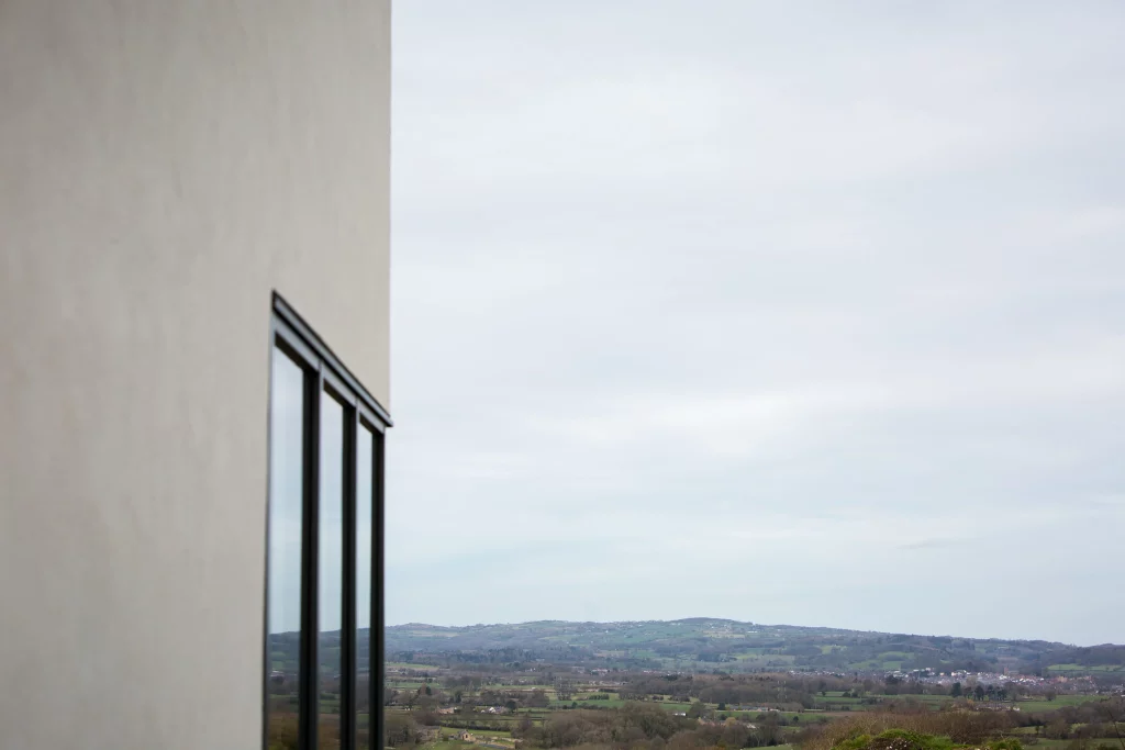 Corner of a modern beige building with tall glass windows overlooking a distant green hilly landscape under a pale sky.