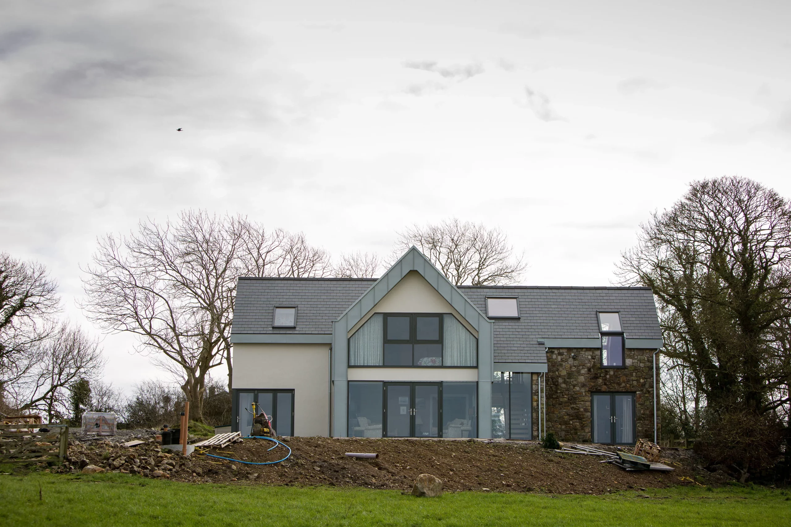Modern two-storey house with large central glass doors and gabled roof, bare trees around and a muddy foreground with construction materials.