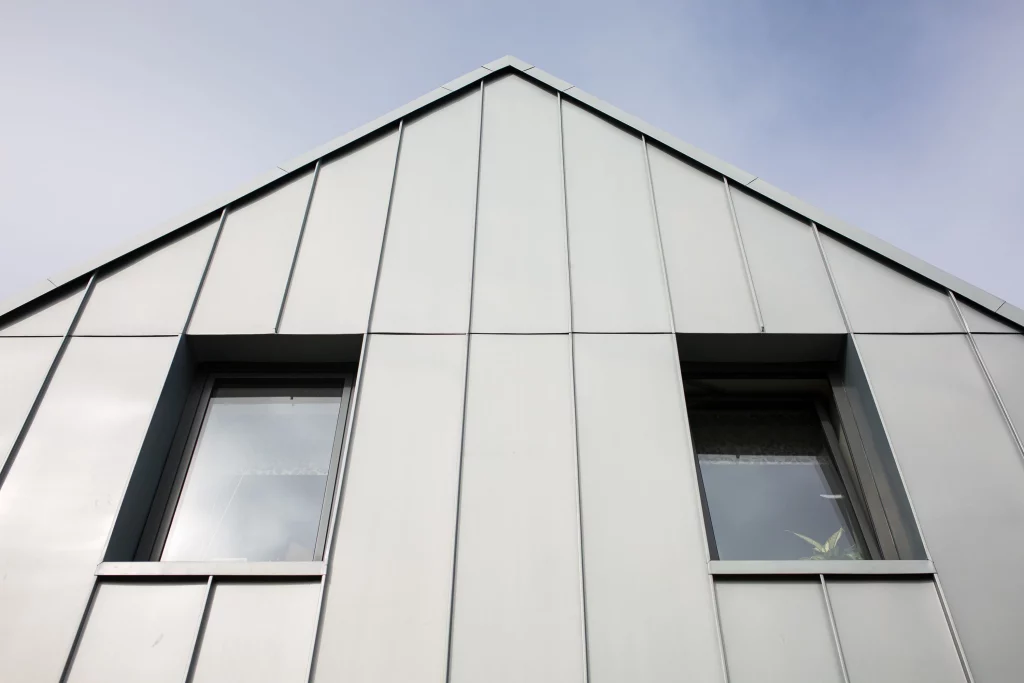 Gabled modern building façade clad in vertical metal panels with two narrow recessed windows under a pale sky