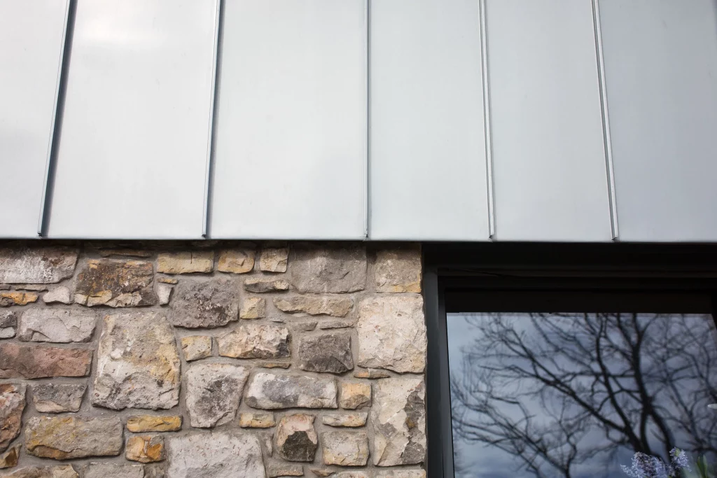 Stone exterior wall with irregular beige blocks under a pale metal awning and a dark-framed window reflecting tree branches