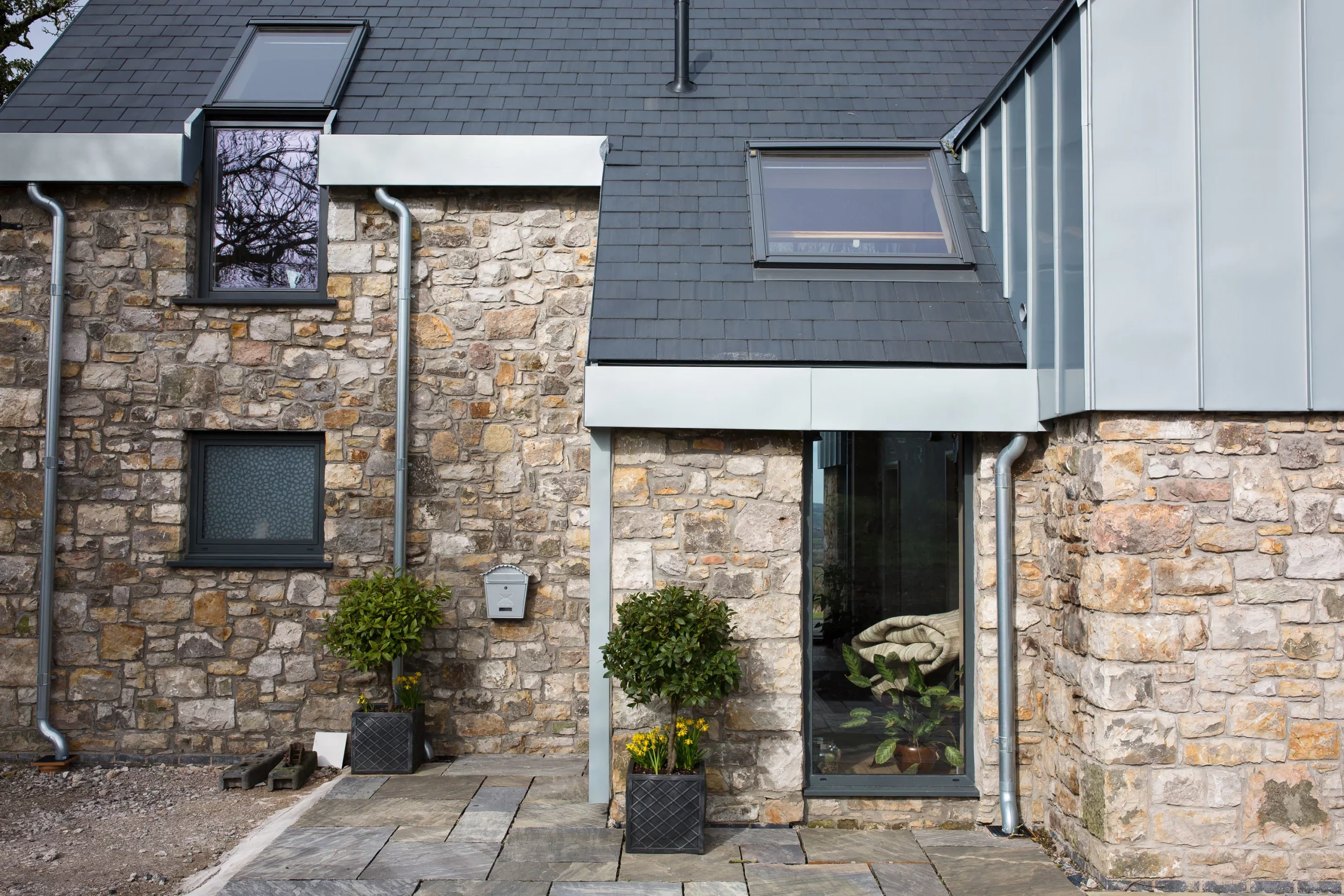 Stone cottage exterior with slate roof, two skylight windows, rain gutters and potted shrubs by a glass door.