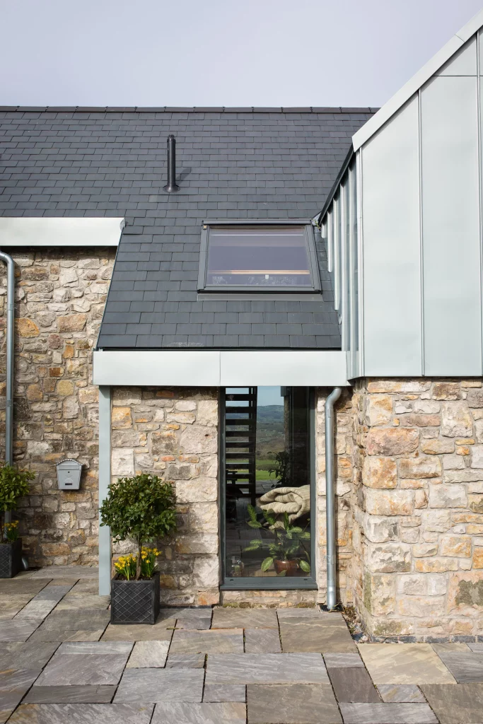 Stone cottage wall with slate roof, small dormer window and narrow glass door flanked by potted topiary and daffodils