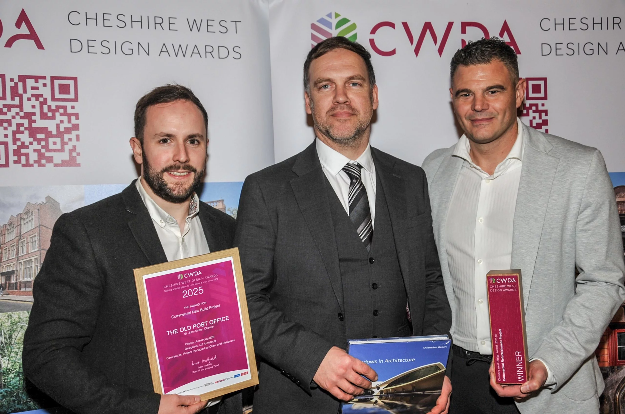 Three men in suits standing side by side at an awards backdrop, the left man holding a framed 2025 Cheshire West Design Awards certificate and the right man holding a red winner trophy.