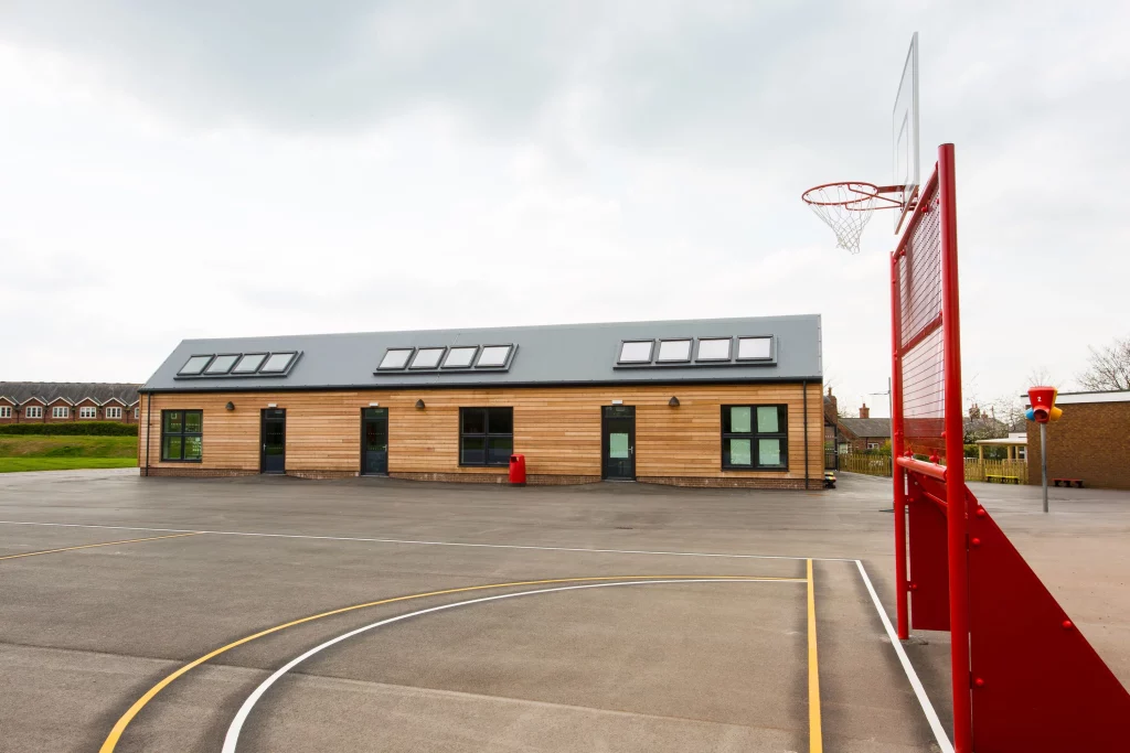 Empty school playground with painted court lines, wooden single-storey building and a red basketball hoop on the right.