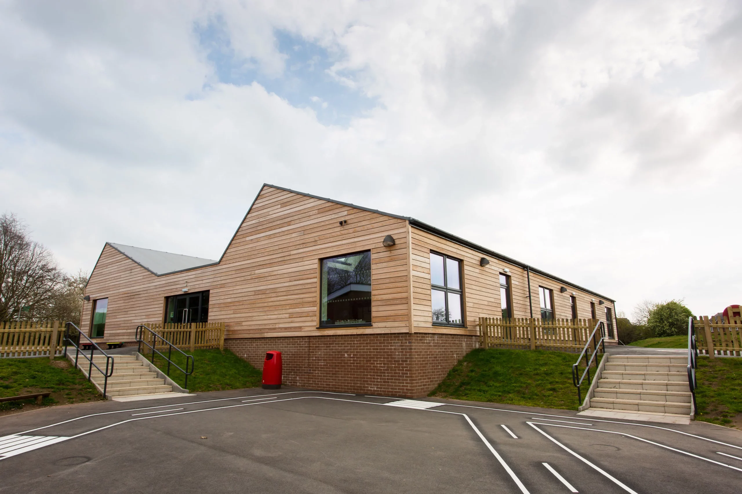 Single-storey modern wooden school building with large windows, two stairways and a playground road marking in foreground.