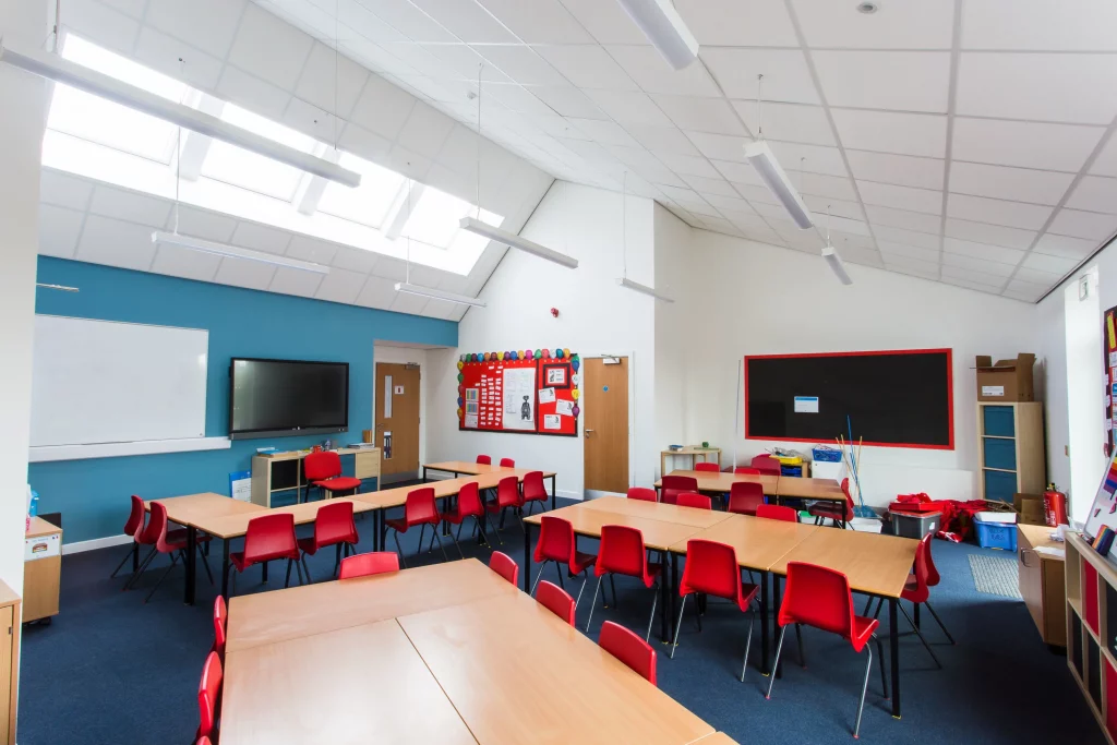 Classroom with rows of wooden tables and red chairs, skylights and hanging lights, whiteboard, bulletin boards and storage units