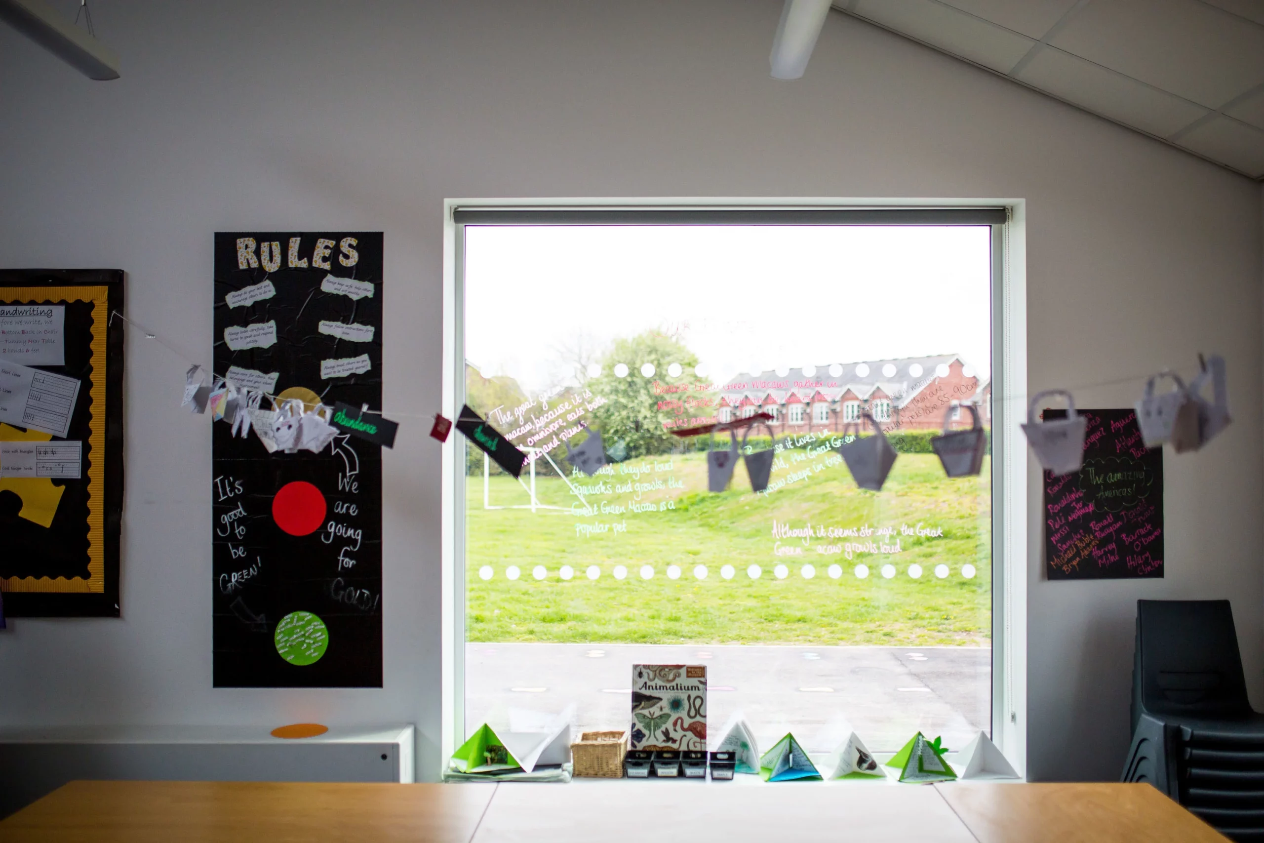 Classroom window with paper craft garlands, a "RULES" poster on the left and stacked chairs on the right, view of grassy playground outside.