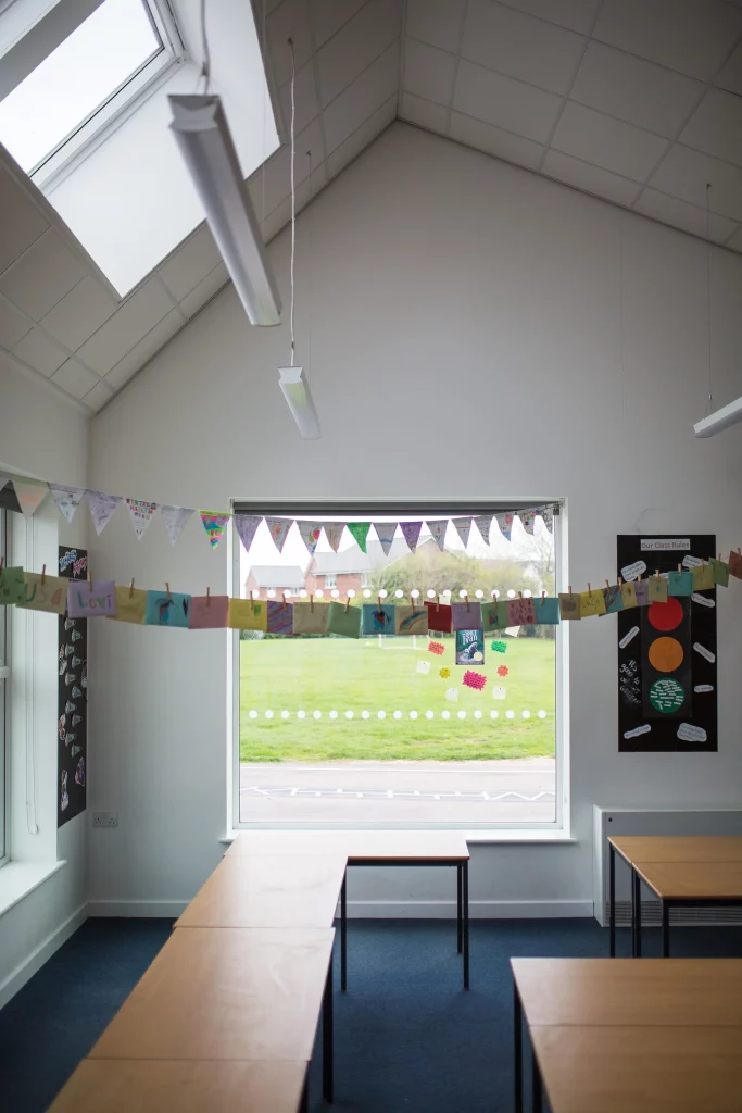 Classroom interior with hanging bunting and student artwork across a large window, rows of desks and suspended ceiling lights