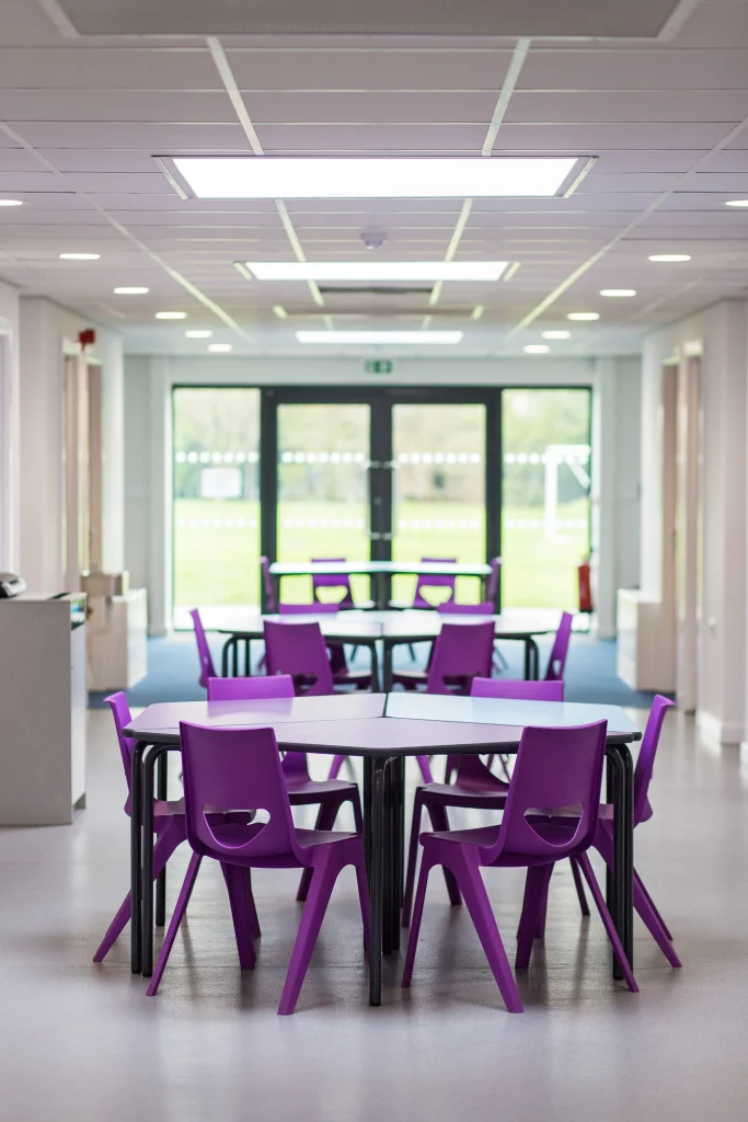 Empty school dining area with purple plastic chairs around hexagonal tables and double doors at the far end of the corridor