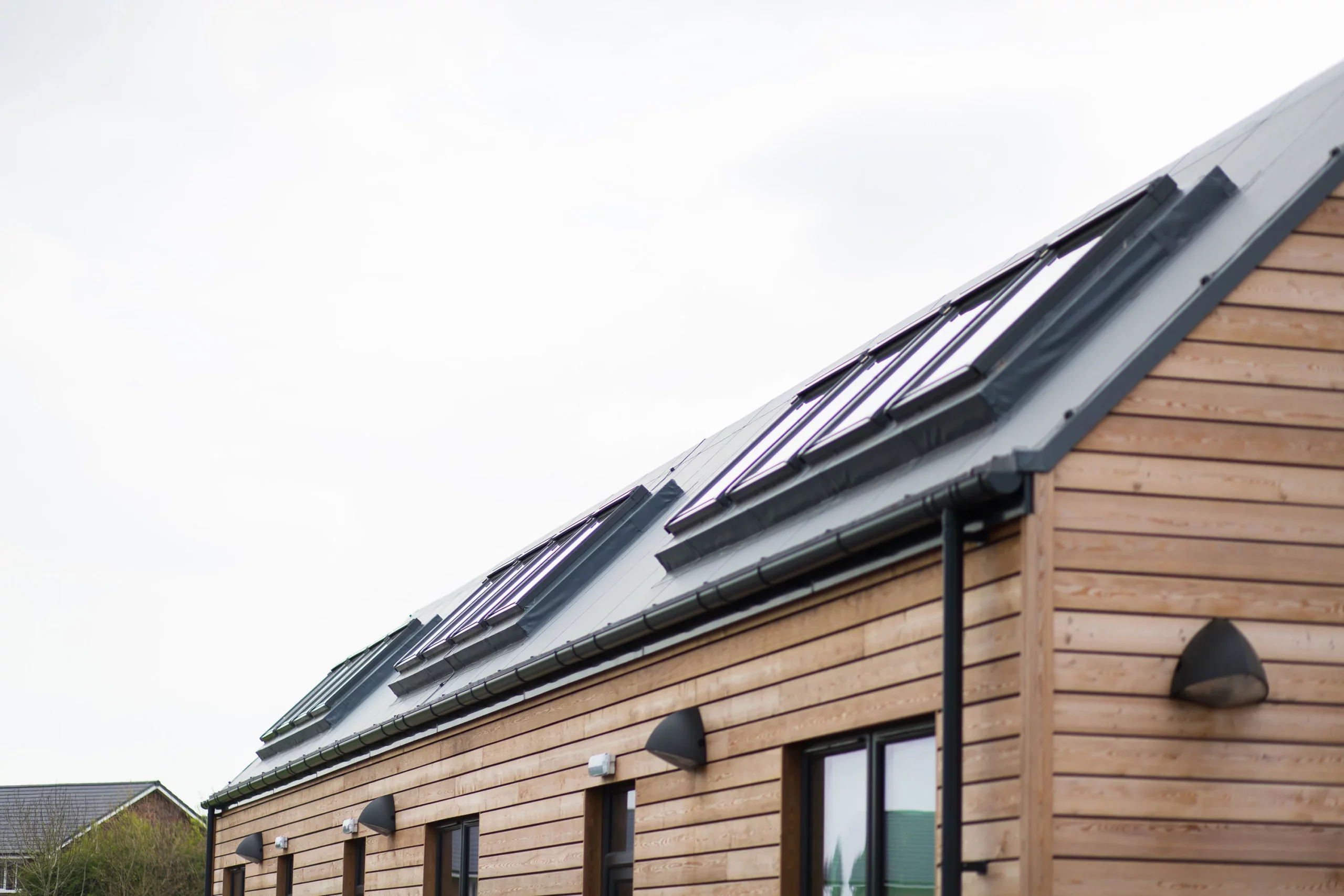 Row of modern wooden houses with sloped metal roofs and multiple roof windows under an overcast sky