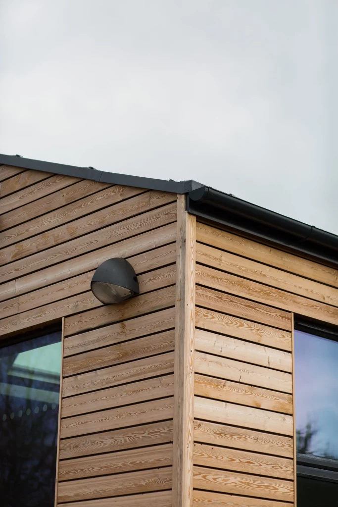 Corner of a modern wooden-clad building with horizontal planks, black guttering and a wall-mounted round exterior light.