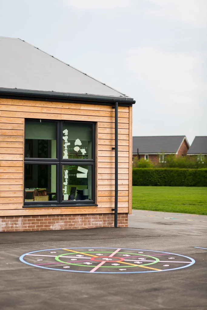 Corner of a modern wooden-clad school building with black-framed windows and a colourful painted hopscotch circle on the playground