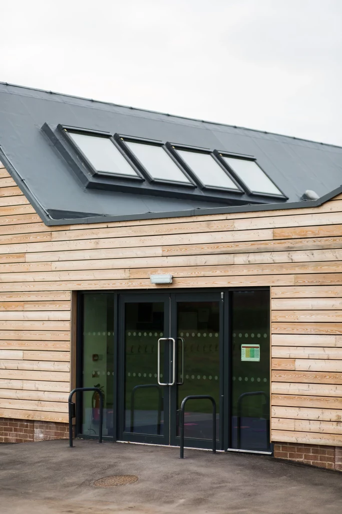 Modern wooden building entrance with glass double doors and three angled skylights on a sloped dark roof