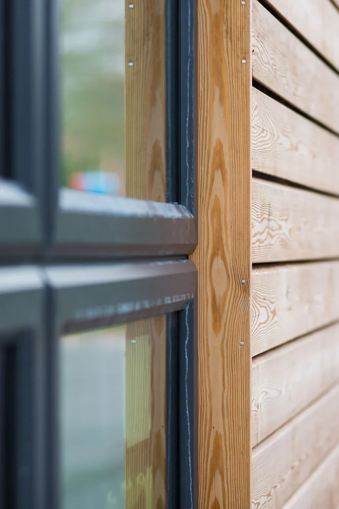 Close-up of a wooden exterior wall with horizontal cladding and a black metal-framed window trim in soft focus.