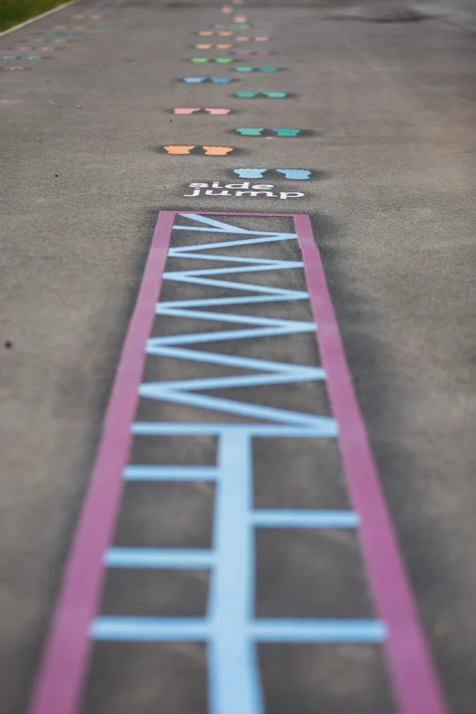 Painted playground path on pavement with colourful ladder and footprint hop markers and the words "side jump" visible.