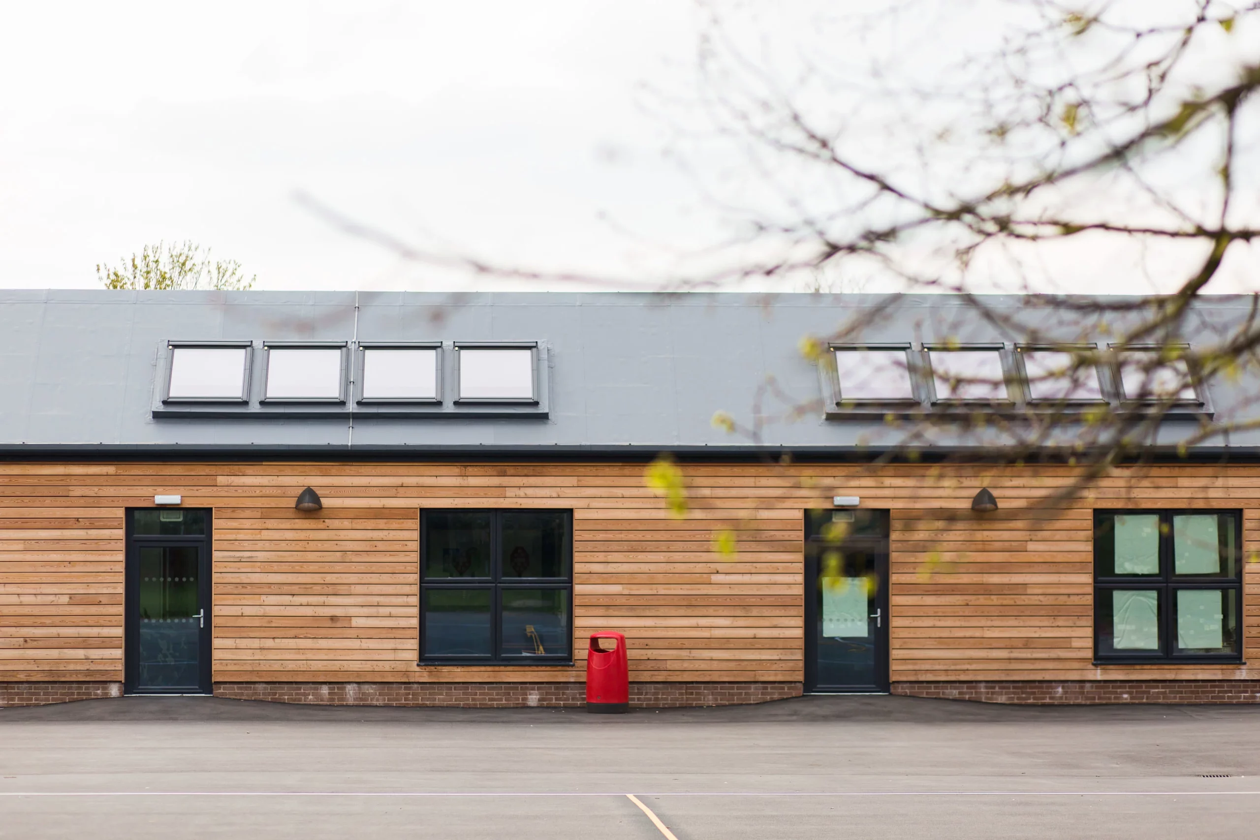 Single-storey building with horizontal wooden cladding, two dark doors and windows, a row of roof skylights and a red bin outside.
