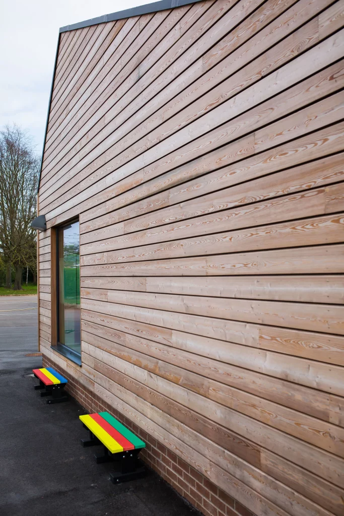 Angled view of a modern building clad in horizontal light-wood panels with two small rainbow-painted benches against a brick plinth.