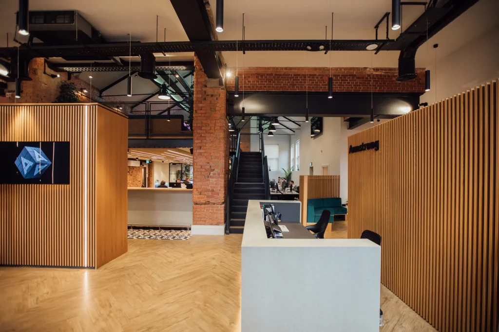 Modern open-plan office reception with wooden slat panels, central staircase, exposed brick and ceiling beams.