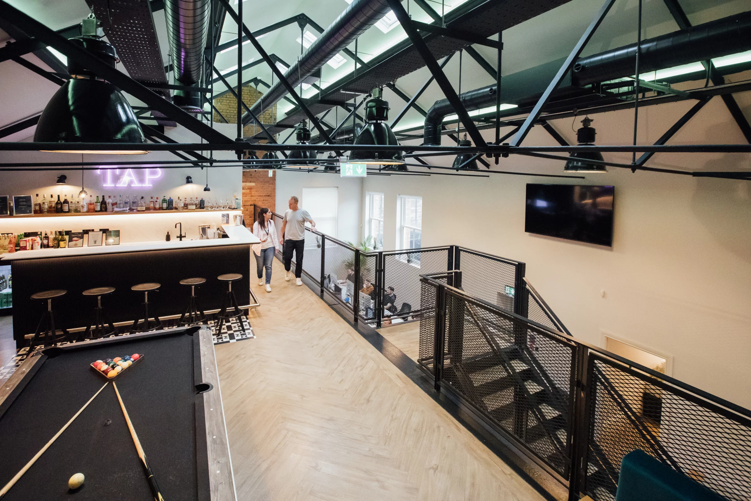 Open-plan loft lounge with pool table, bar counter and two people walking past a metal-railed mezzanine staircase.
