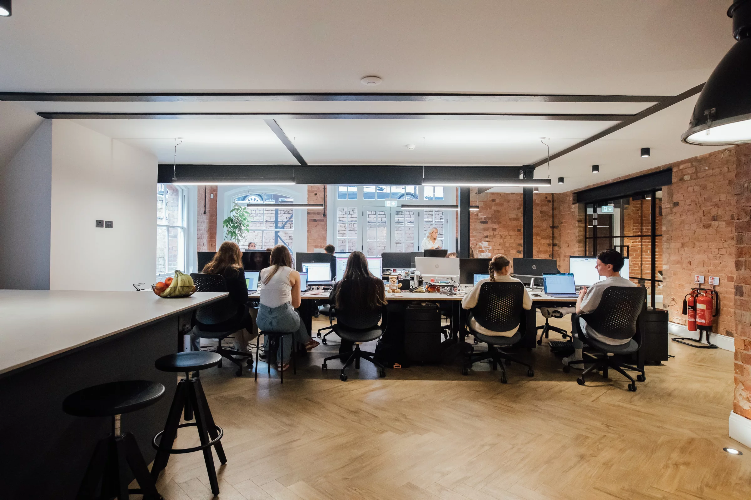 Open-plan office with rows of people working at desks, exposed brick walls, hanging lights and wooden herringbone floor