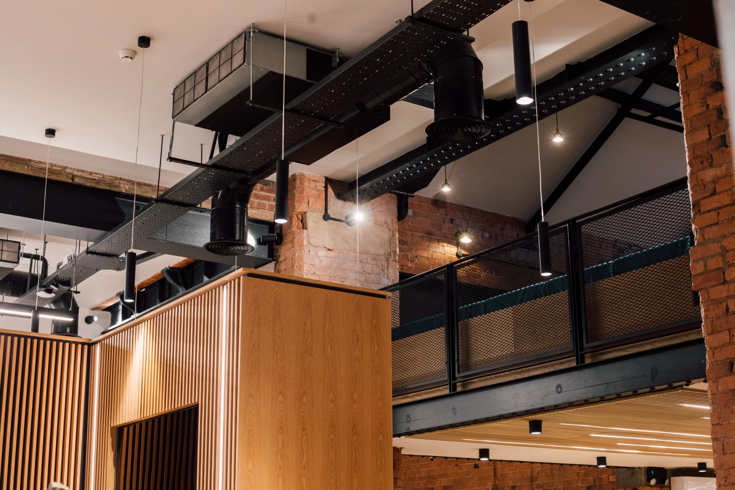 Exposed industrial ceiling with black ductwork, hanging cylinder pendant lights and wooden slatted partition below