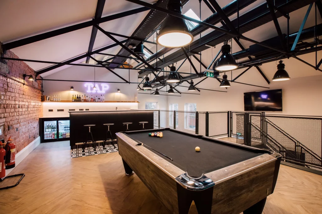 Loft-style games room with a black pool table, overhead industrial lights, bar counter with stools and neon "TAP" sign.