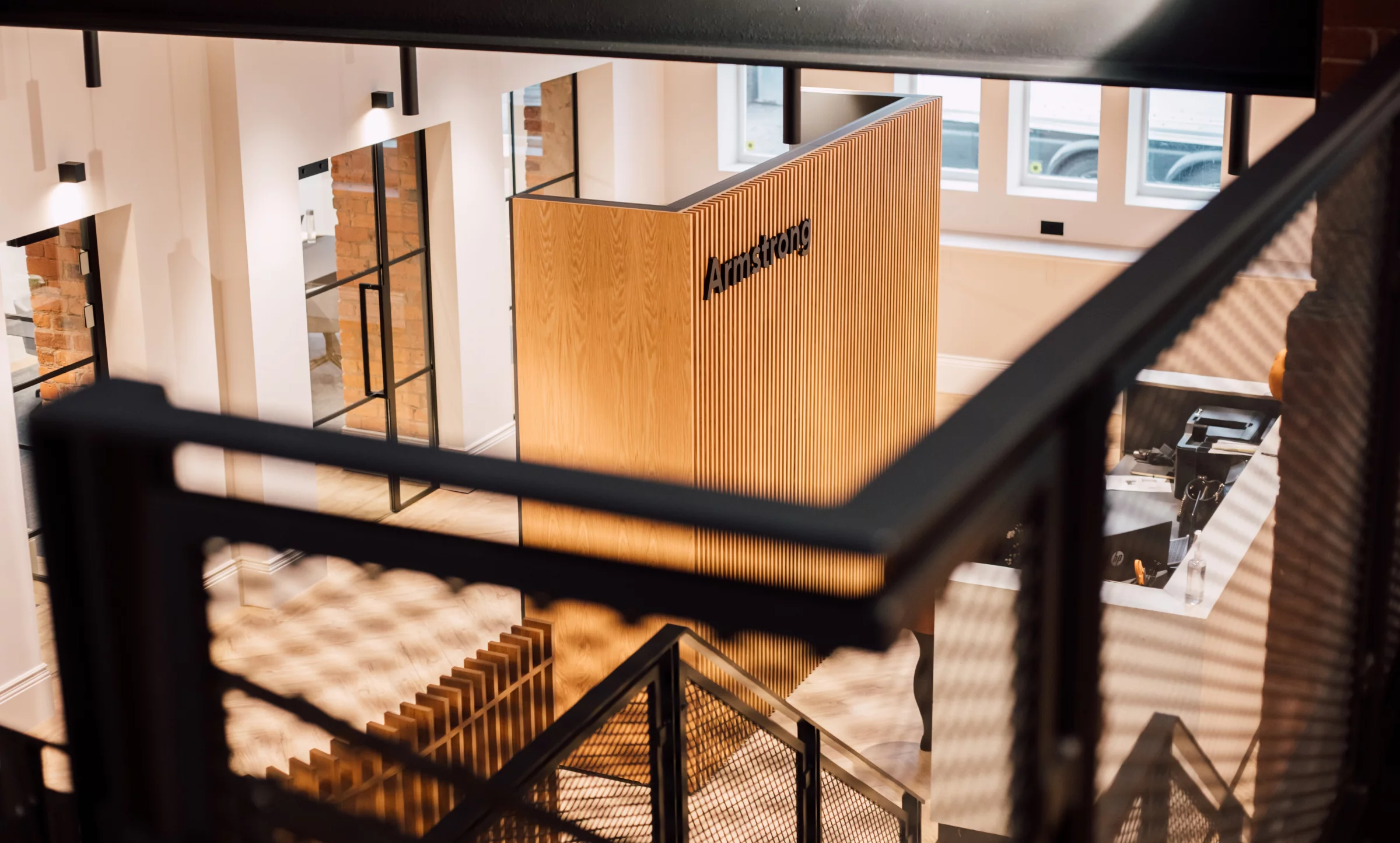 View down into a modern office with a tall slatted oak reception wall labelled "Armstrong", black metal stair railings and glass-partitioned rooms.