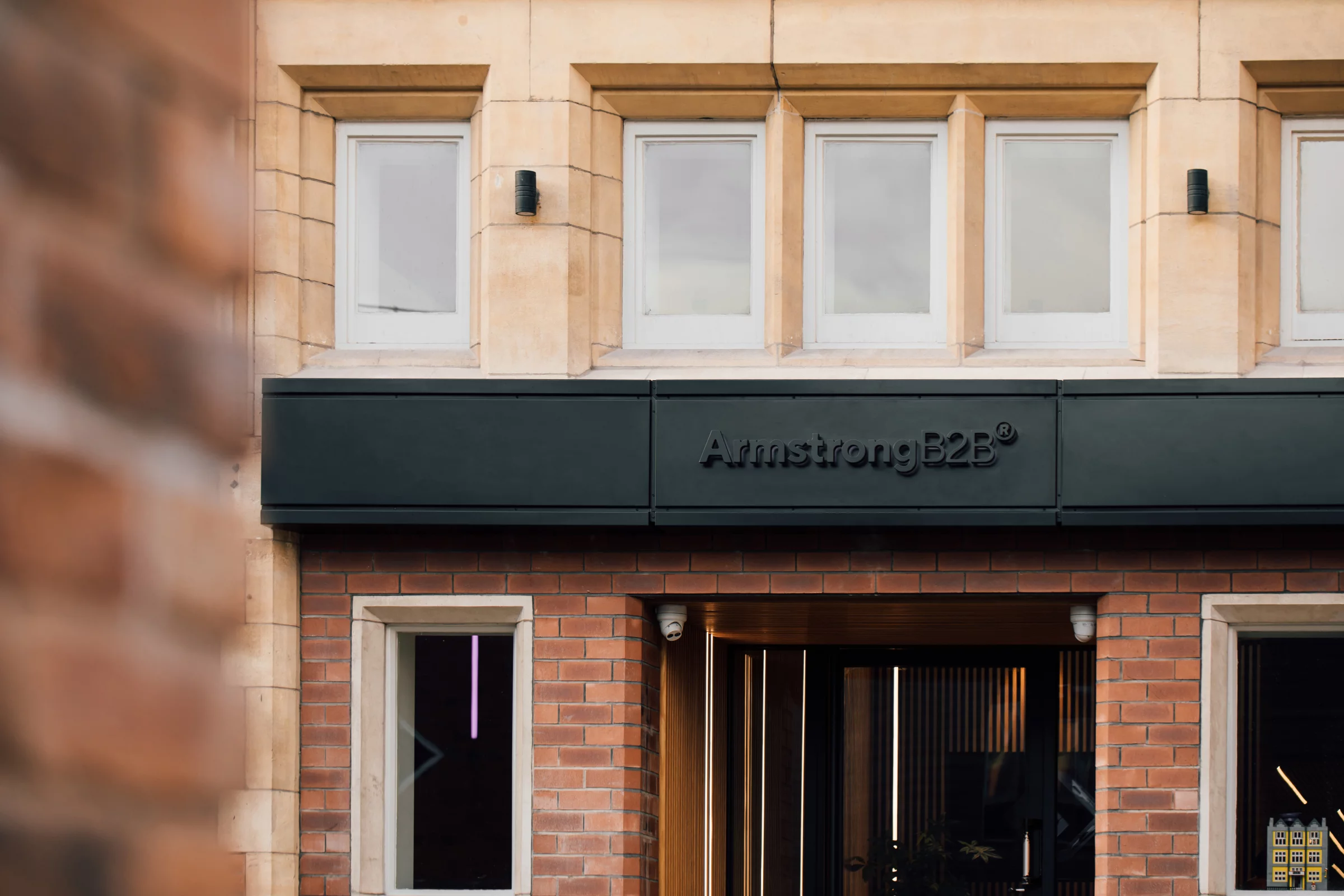Street-level view of a brick and stone storefront with black sign reading "ArmstrongB2B" and upper white-framed windows