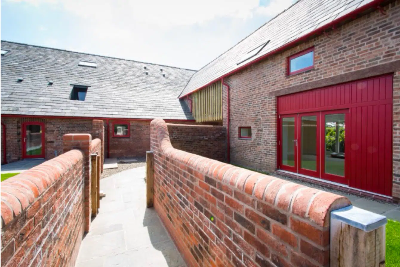 Walled brick courtyard with a curved brick walkway leading to modern barn-style buildings with red-framed doors and windows.