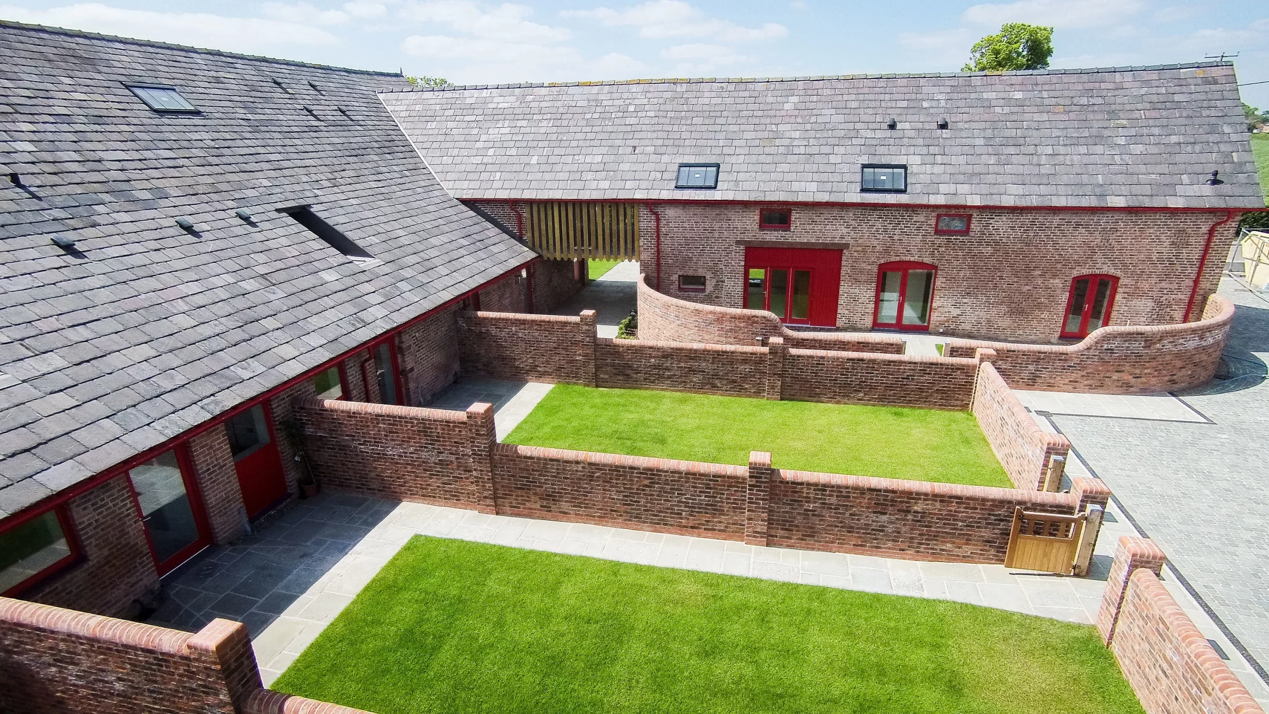 Courtyard of converted red-brick barns with slate roofs, red doors and windows, paved paths and small enclosed lawns.