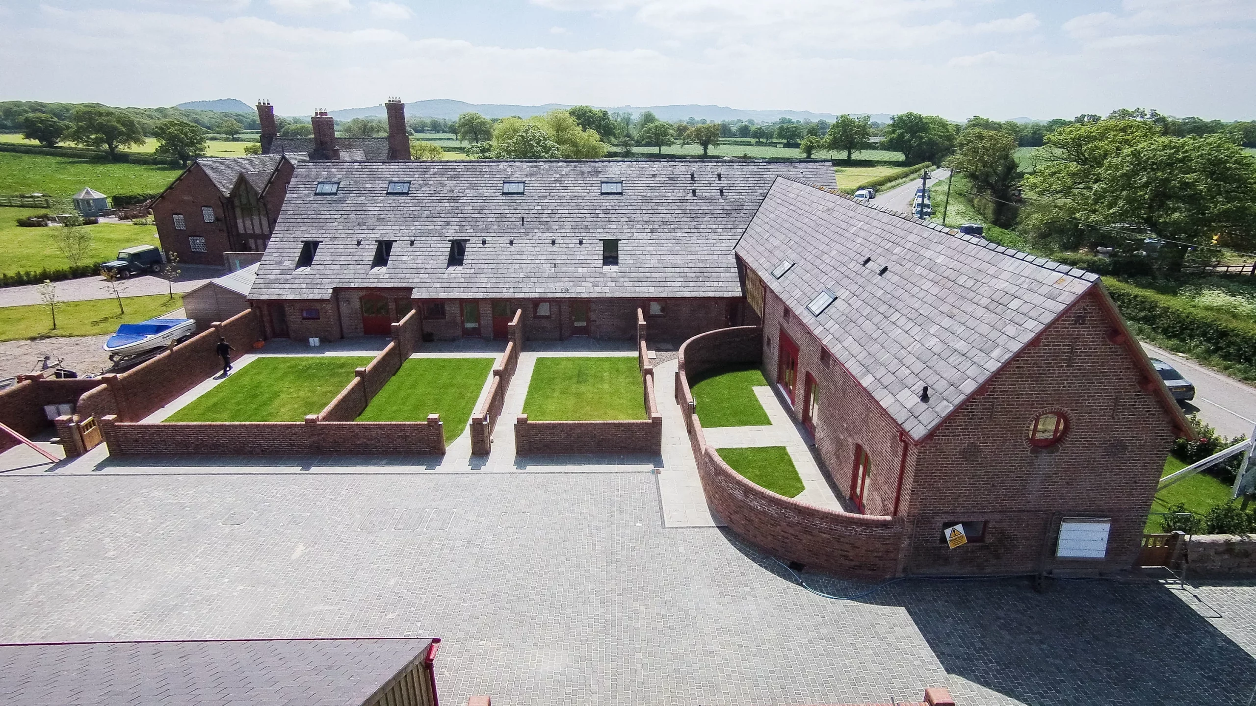 Aerial view of a red-brick courtyard with three small enclosed lawns, slate roofs and surrounding countryside.
