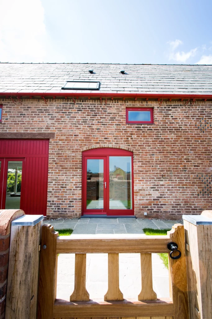 Front view of a red-framed glass door set in a brick house with slate roof, small red window above and wooden gate in foreground