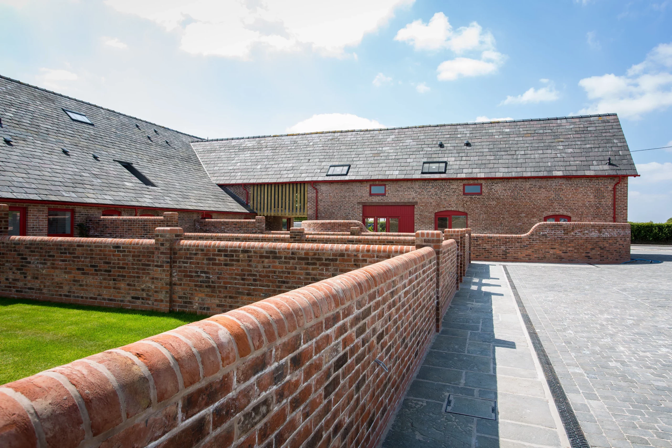 Long red-brick courtyard wall leading to a renovated brick barn with slate roofs, skylights and paved driveway under blue sky