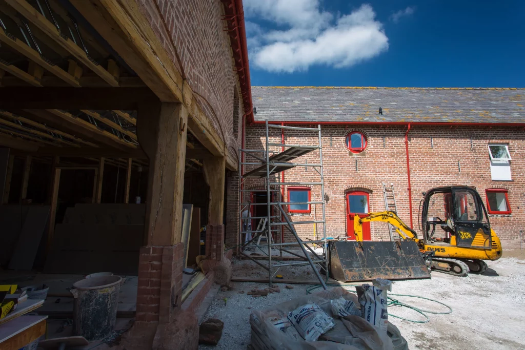 Renovation site with exposed timber framing, brick building with red-trimmed windows and doors, scaffolding and a yellow JCB digger.