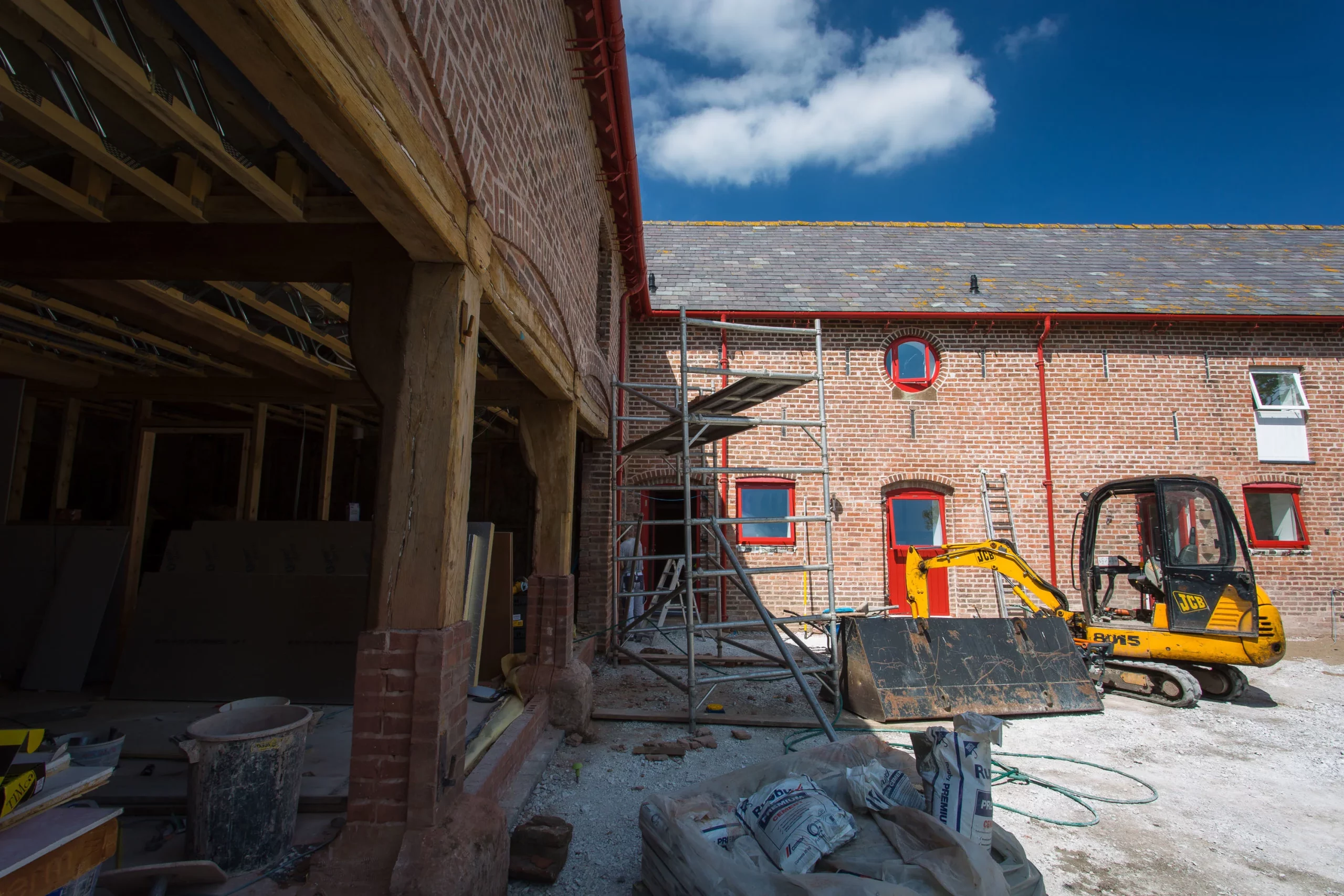 Renovation site with exposed timber framing, brick building with red-trimmed windows and doors, scaffolding and a yellow JCB digger.