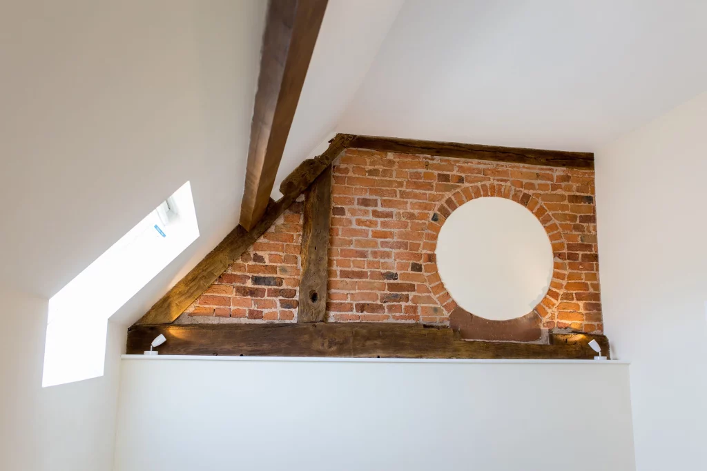 Exposed red brick wall with wooden beams and a circular plaster patch beneath a sloped white ceiling and skylight