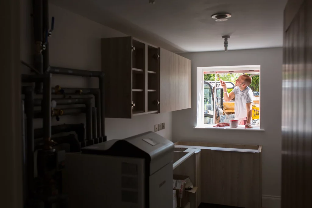 Man in a t-shirt painting the window trim from an open window in a dimly lit utility room with exposed pipes and cabinets.