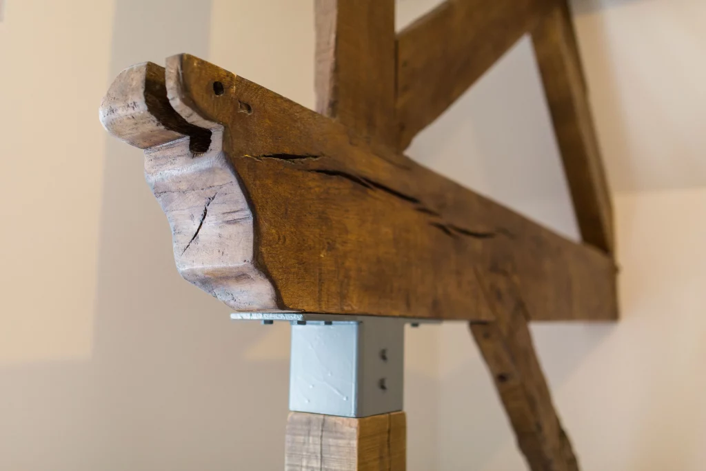 Close-up of a cracked, carved wooden beam supported on a painted metal bracket, showing wood grain and join details.