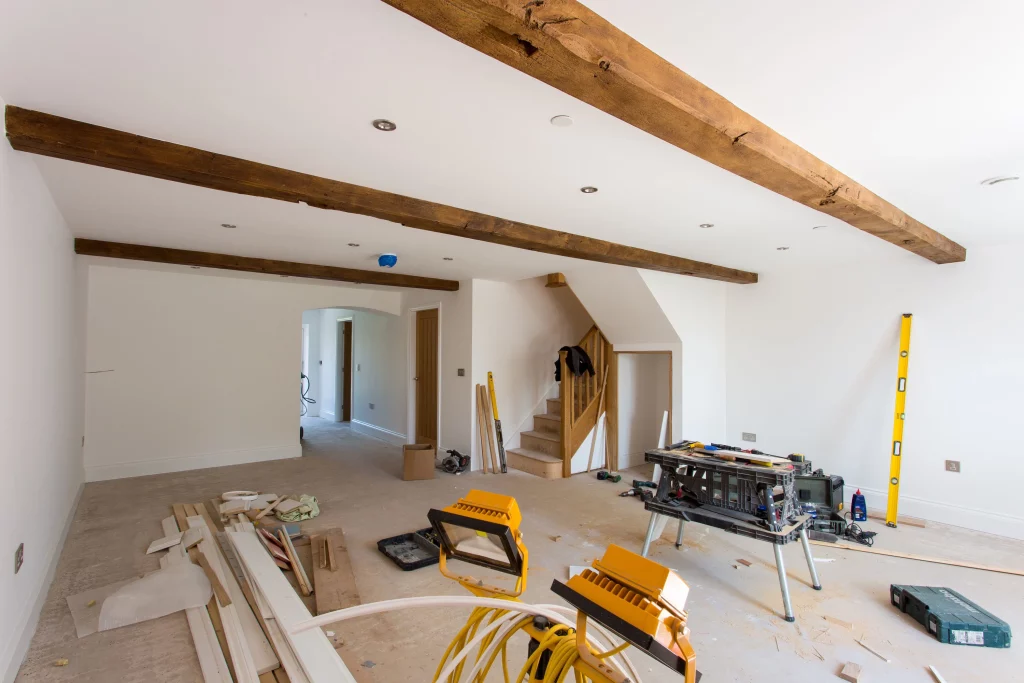 Unfurnished room under renovation with exposed wooden ceiling beams, construction tools, lumber and a stepladder scattered on the floor