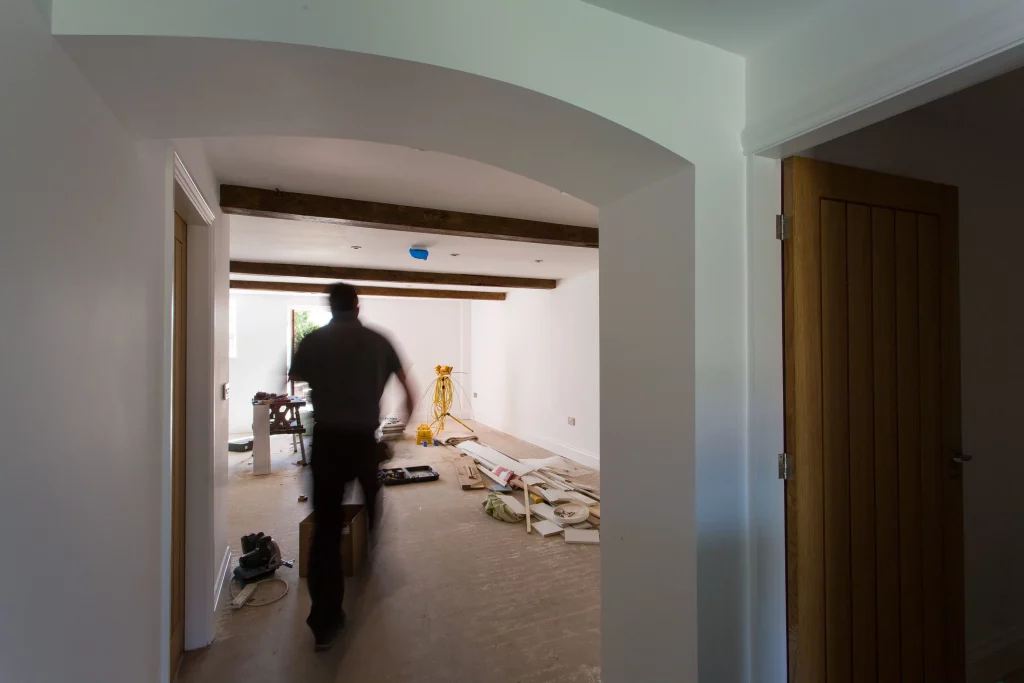 Blurry contractor walking through a white-walled room under renovation with exposed wooden beams and scattered tools and materials.