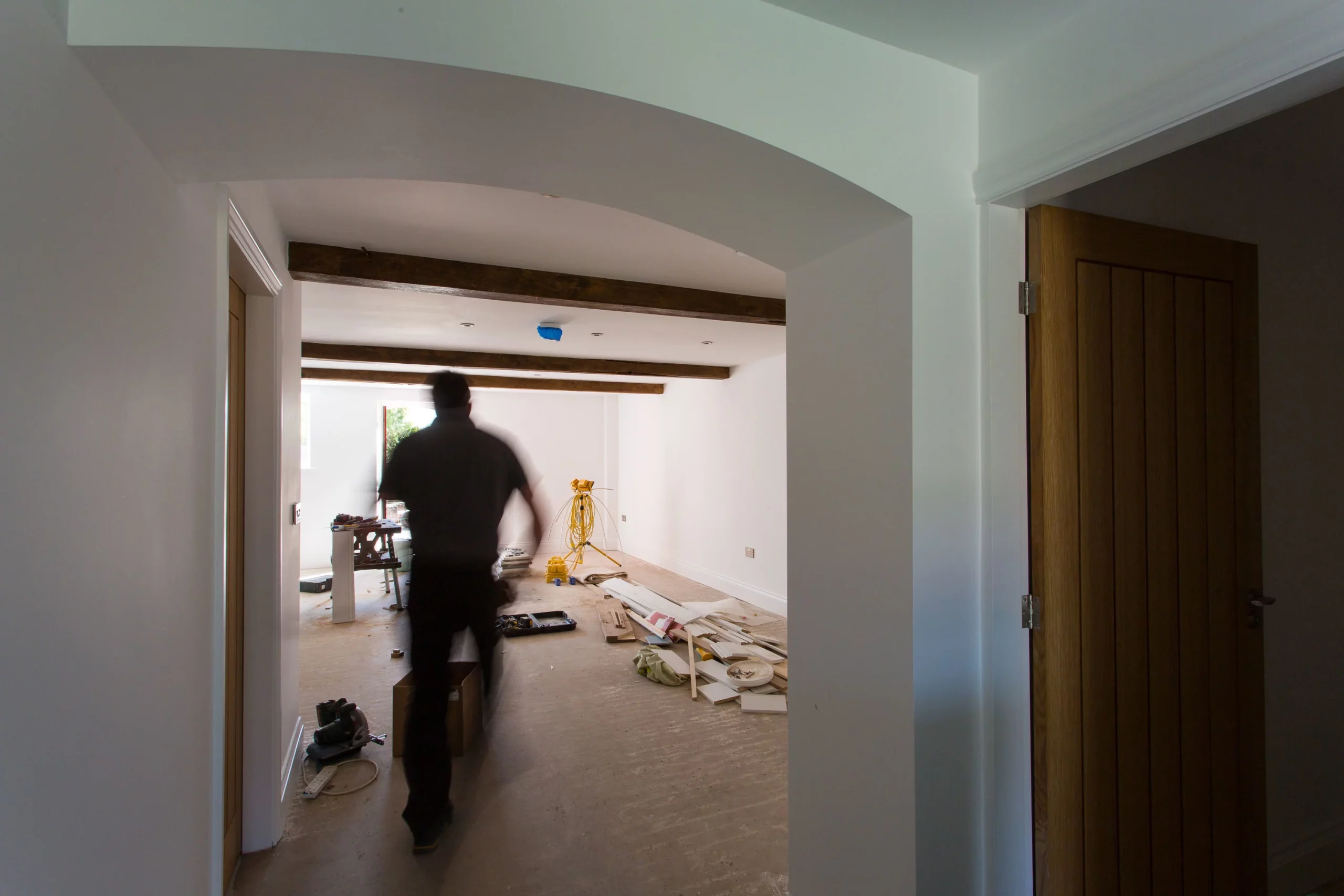 Blurry contractor walking through a white-walled room under renovation with exposed wooden beams and scattered tools and materials.