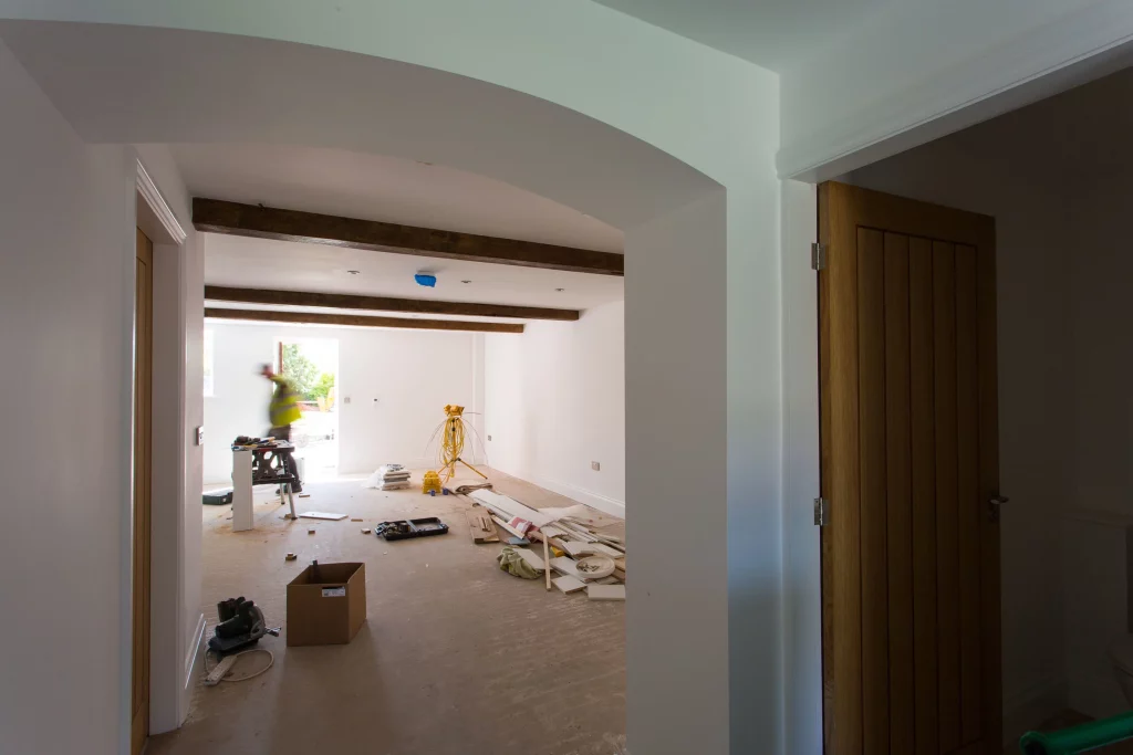Open-plan room under renovation with exposed ceiling beams, tools, scattered building materials and a work light tripod.
