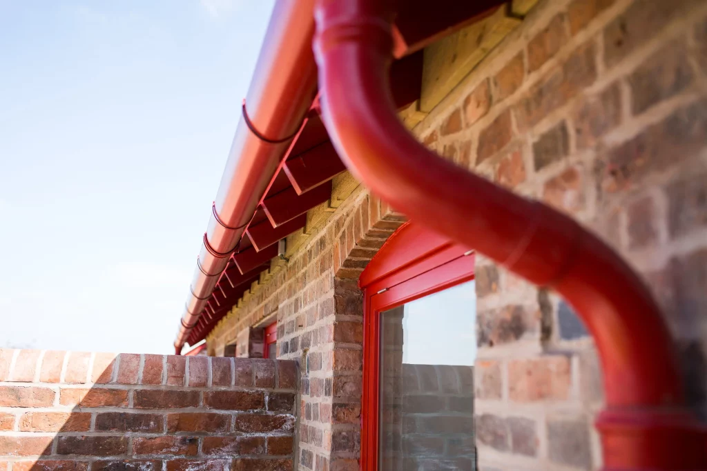 Red painted gutter and downpipe running along the eaves of a brick building with a red window frame.
