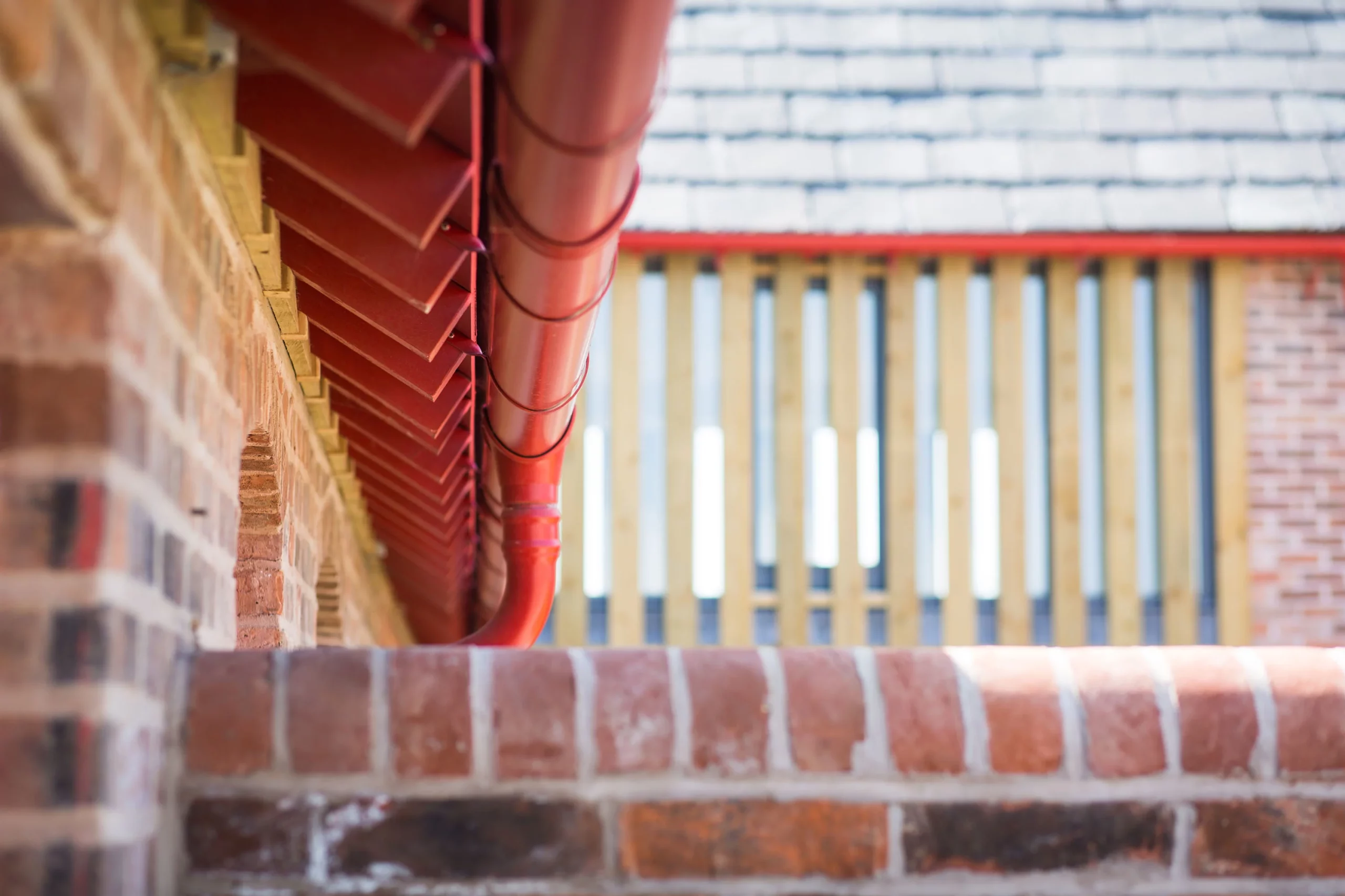 Red metal gutter and downpipe running along a brick wall with blurred wooden slats and tiled roof in background