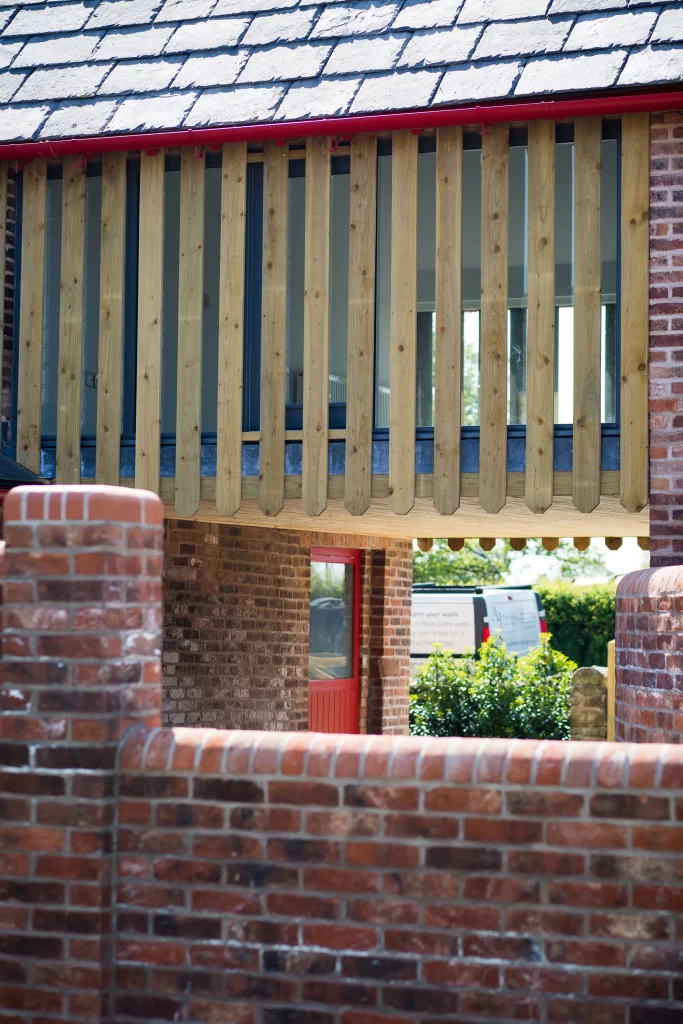 Brick building with a balcony screened by vertical wooden slats, slate roof and a red door visible beneath.