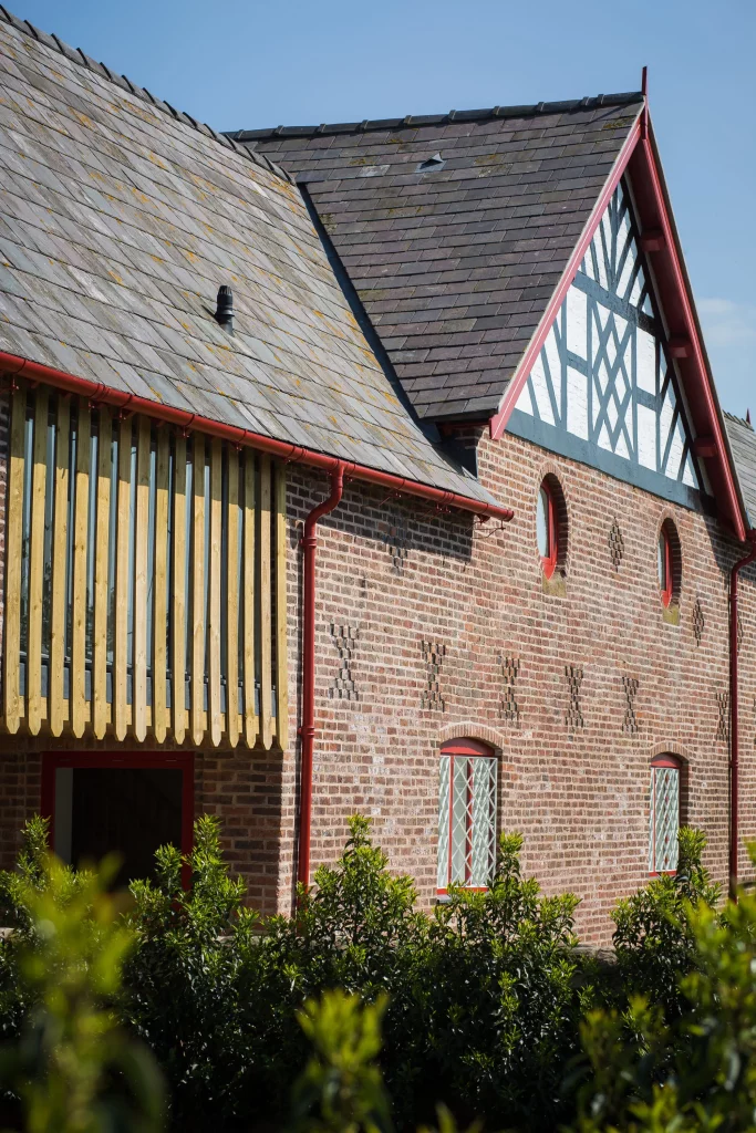 Red-brick Tudor-style building with slate roofs, red trim, lattice windows and a wooden balcony, foreground shrubs.