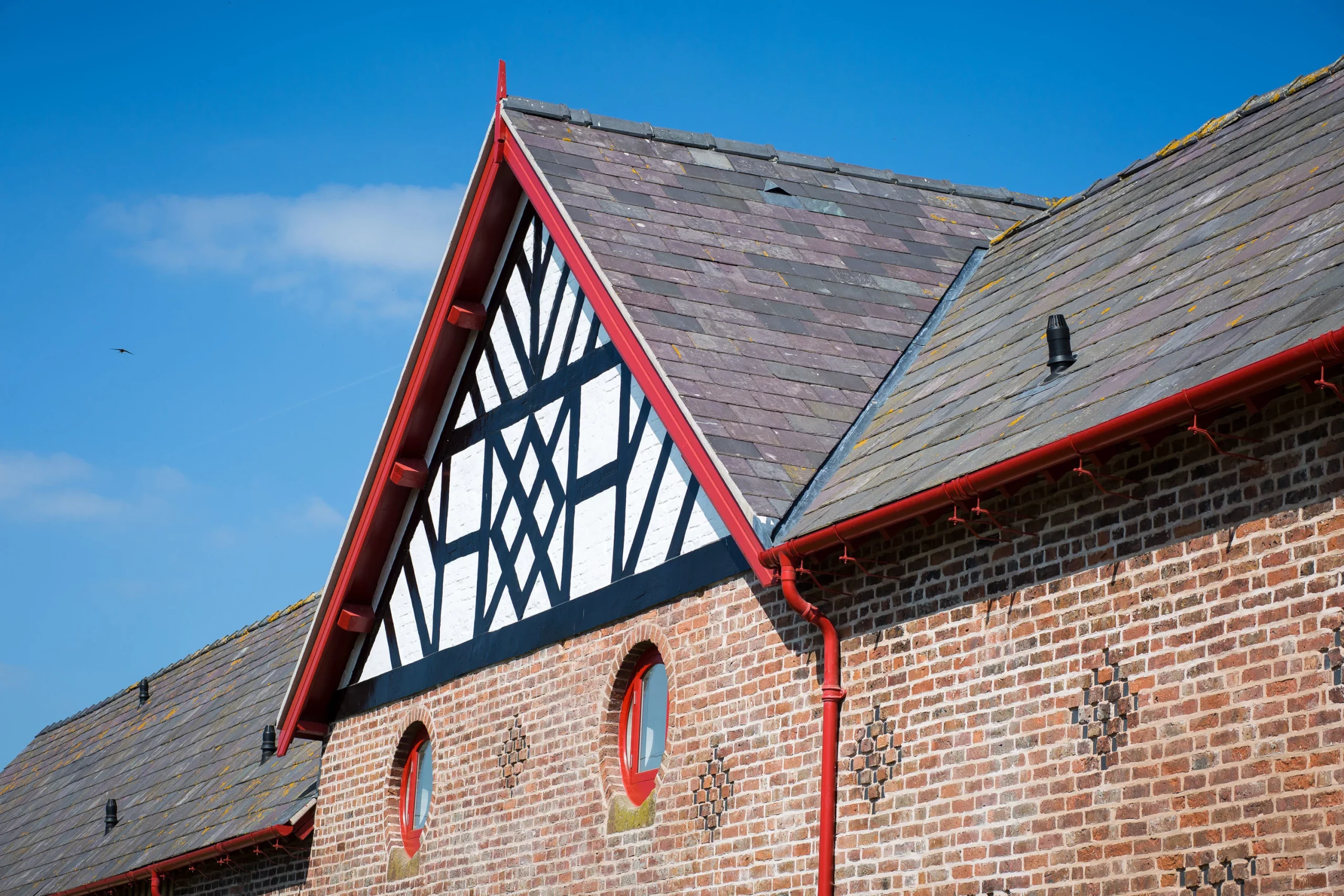 Tudor-style gable with black timber patterns, red trim and round red-framed windows on a brick wall under a slate roof against blue sky