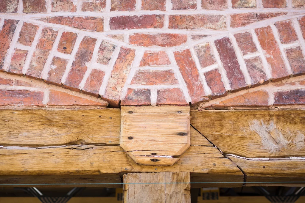 Close-up of a curved redbrick arch with white mortar resting on weathered timber beams and a central wooden corbel.