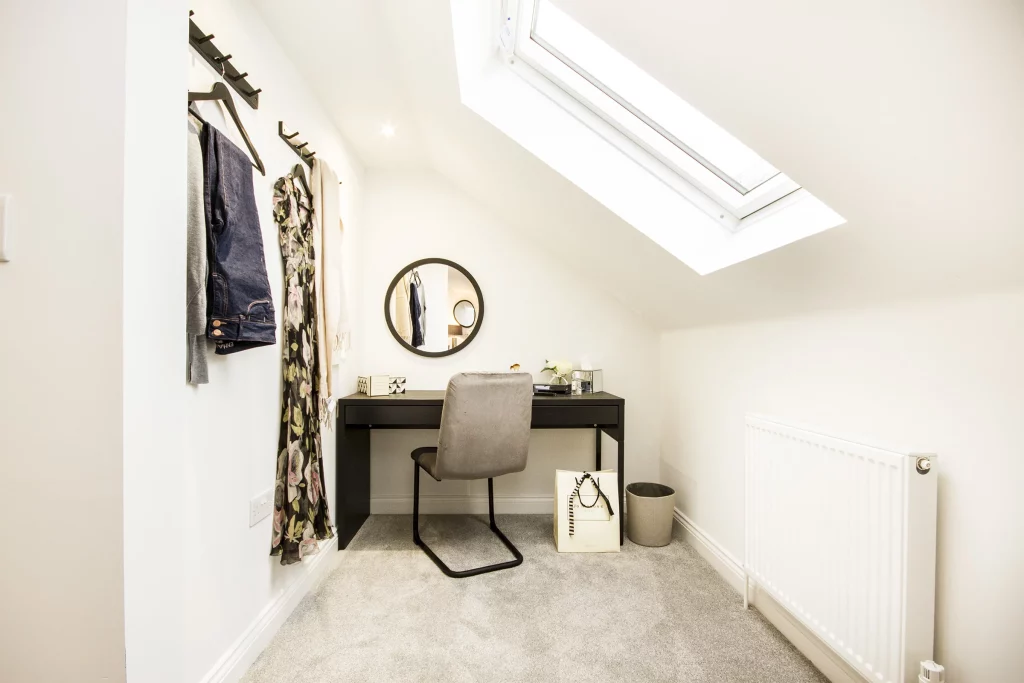 Small bright attic workspace with black desk, grey chair, round wall mirror, skylight and clothes hung on wall hooks